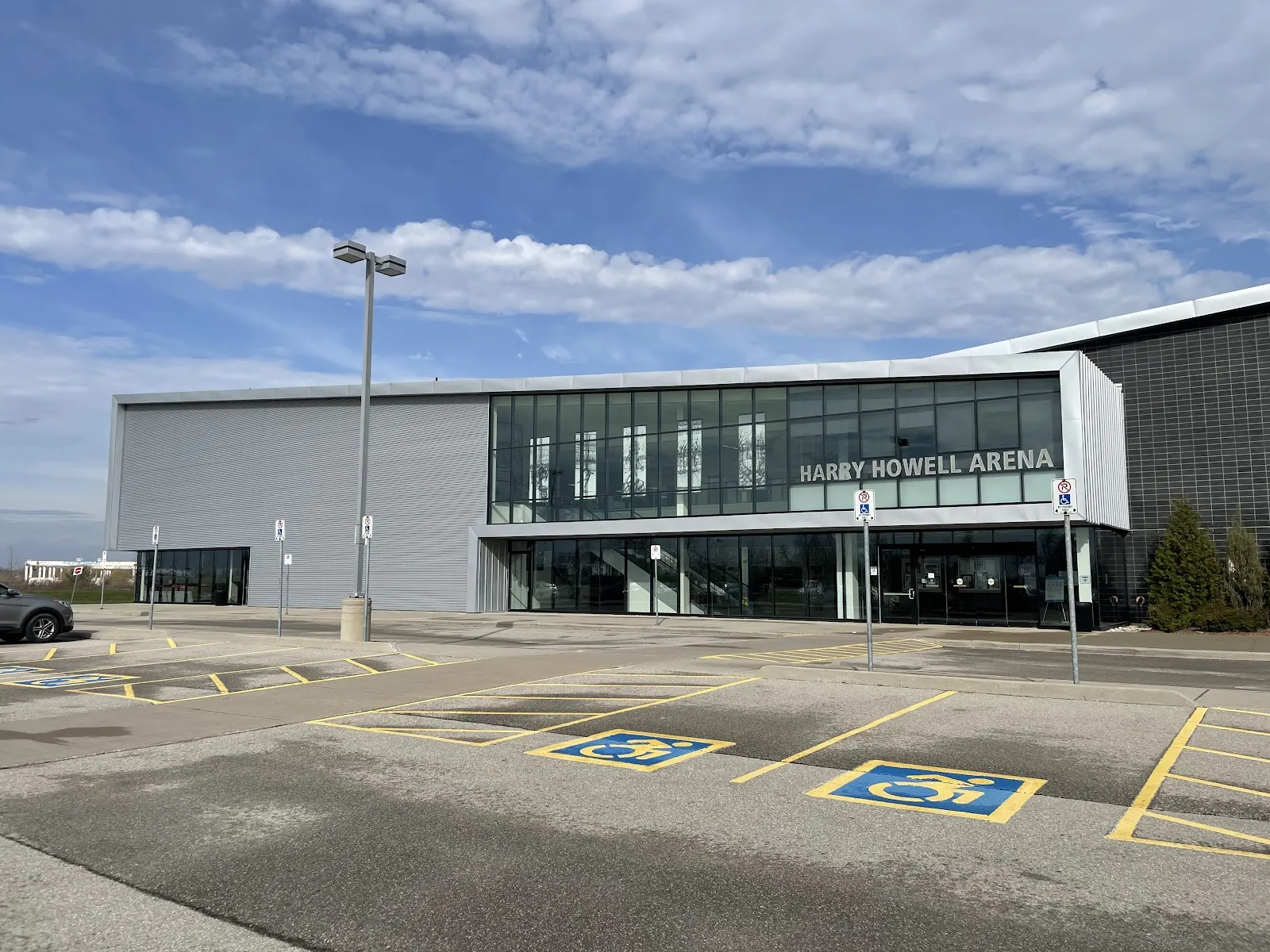 Fachada moderna gris y de vidrio de la arena con la leyenda "Harry Howell Arena", con cajones de estacionamiento accesible al frente. — Harry Howell Twin Pad Arena