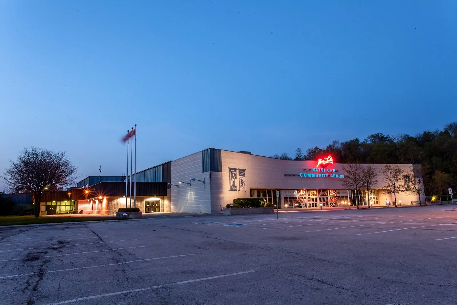 El Harry Lumley Bayshore Community Centre en la hora azul del anochecer, un edificio largo y bajo con un cartel rojo luminoso sobre la entrada, mástiles y árboles a un lado y un aparcamiento vacío delante. — Harry Lumley Bayshore Community Centre