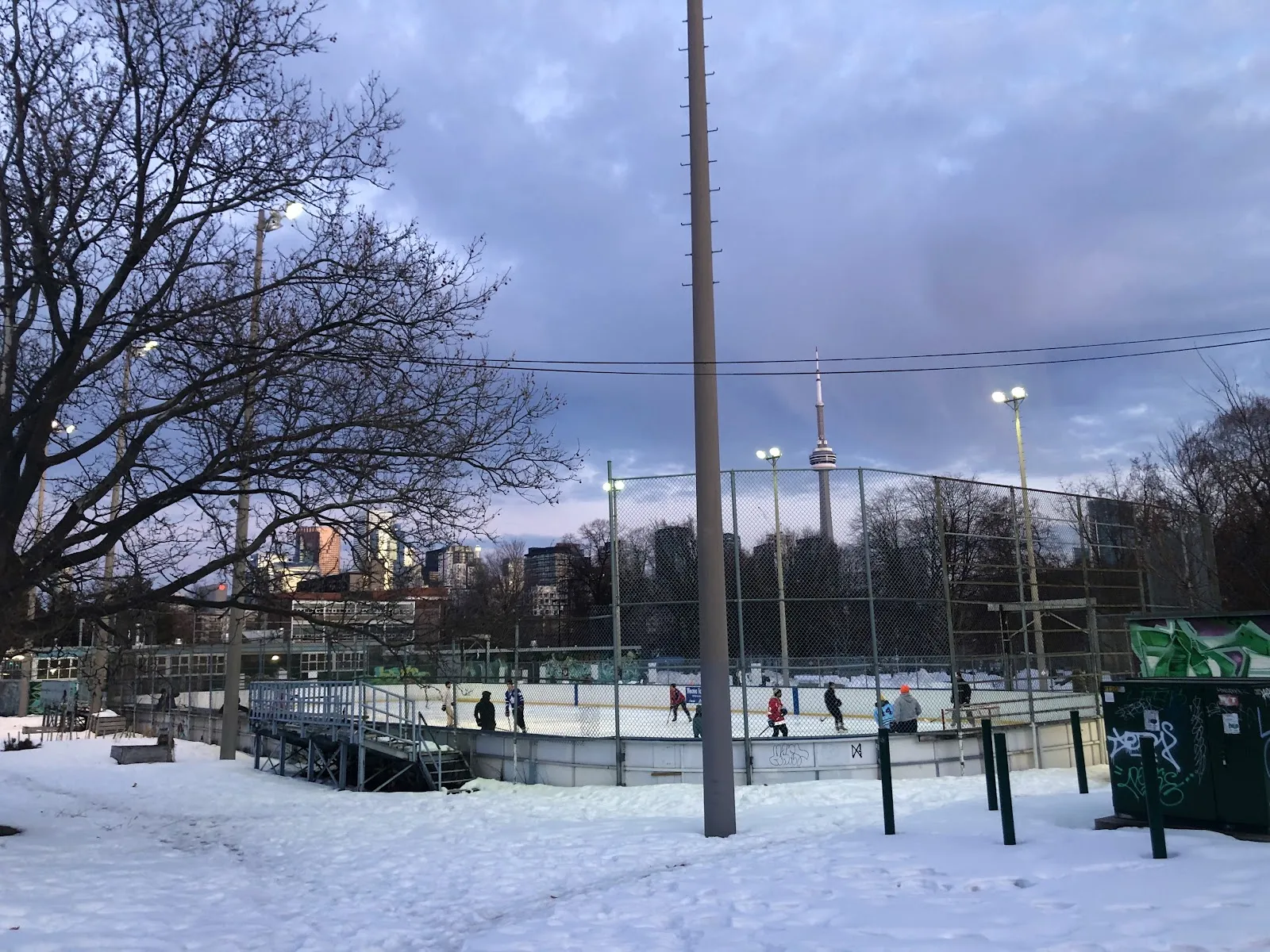 La pista Harry Gairey, cercada e iluminada con reflectores, en Alexandra Park en invierno, con patinadores a lo lejos sobre el hielo y la CN Tower al fondo — Harry R. Gairey Ice Rink (Alexandra Park)