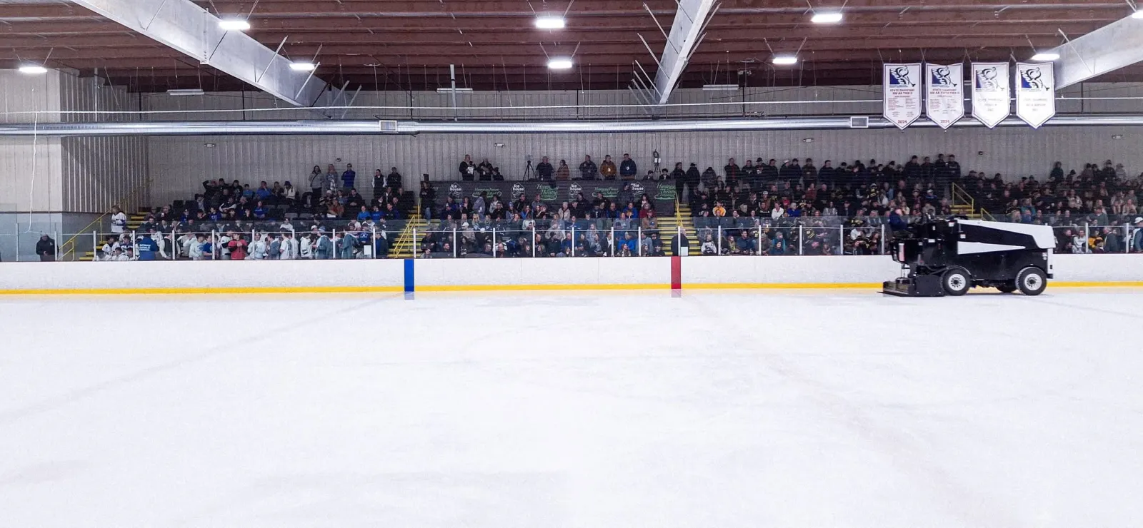 Vista amplia a lo largo de una pista NHL hacia las gradas durante un partido, con una Zamboni estacionada junto a las vallas, mantas de campeonato arriba y las marcas "Ice House" y "Harmony Theatre" visibles en las vallas — Hartland Ice House