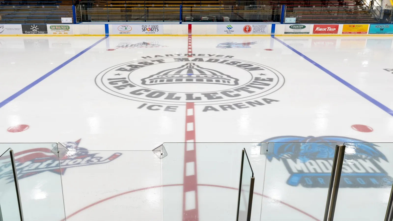 Center ice at Hartmeyer Ice Arena, showing the East Madison Ice Collective center-ice logo, blue lines, dasherboard ad panels and empty bleachers behind the glass. — Hartmeyer Ice Arena