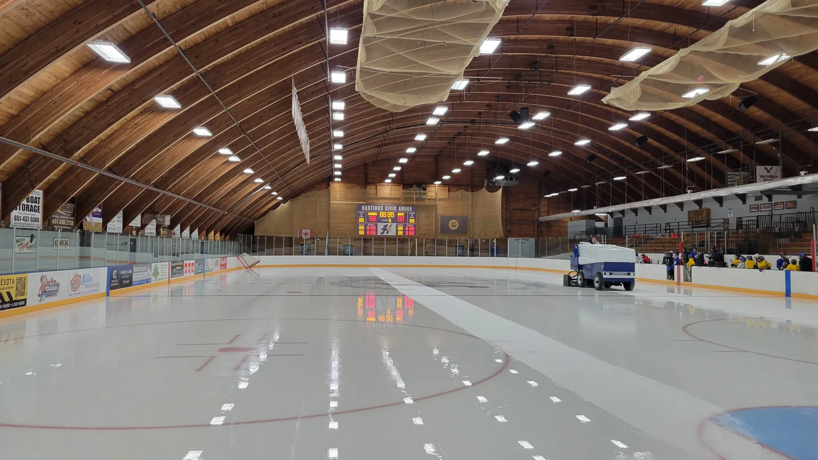 Una Zamboni pule una pista recién inundada bajo el techo curvo de armaduras de madera de la arena, con un marcador que dice "Hastings Civic Arena" al fondo y jugadores reunidos junto a las vallas. — Hastings Civic Arena
