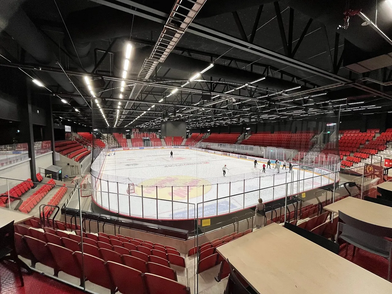 Skaters on the ice of Hatstore Arena seen from the red stands, under the black steel roof — Hatstore Arena
