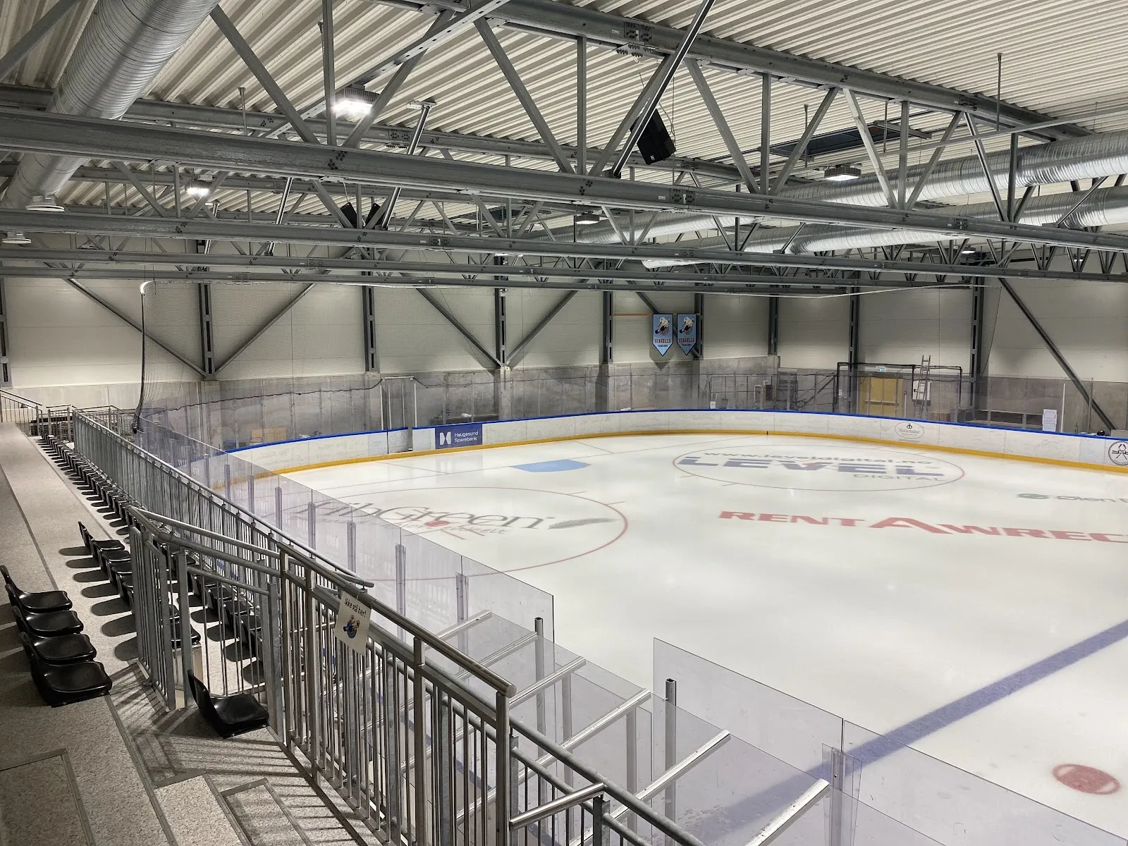 Den tomme isflaten i Haugesund Ishall sett fra tribunen, under et tak med stålsperrer, med vanter og sponsorlogoer på isen. — Haugesund Ishall