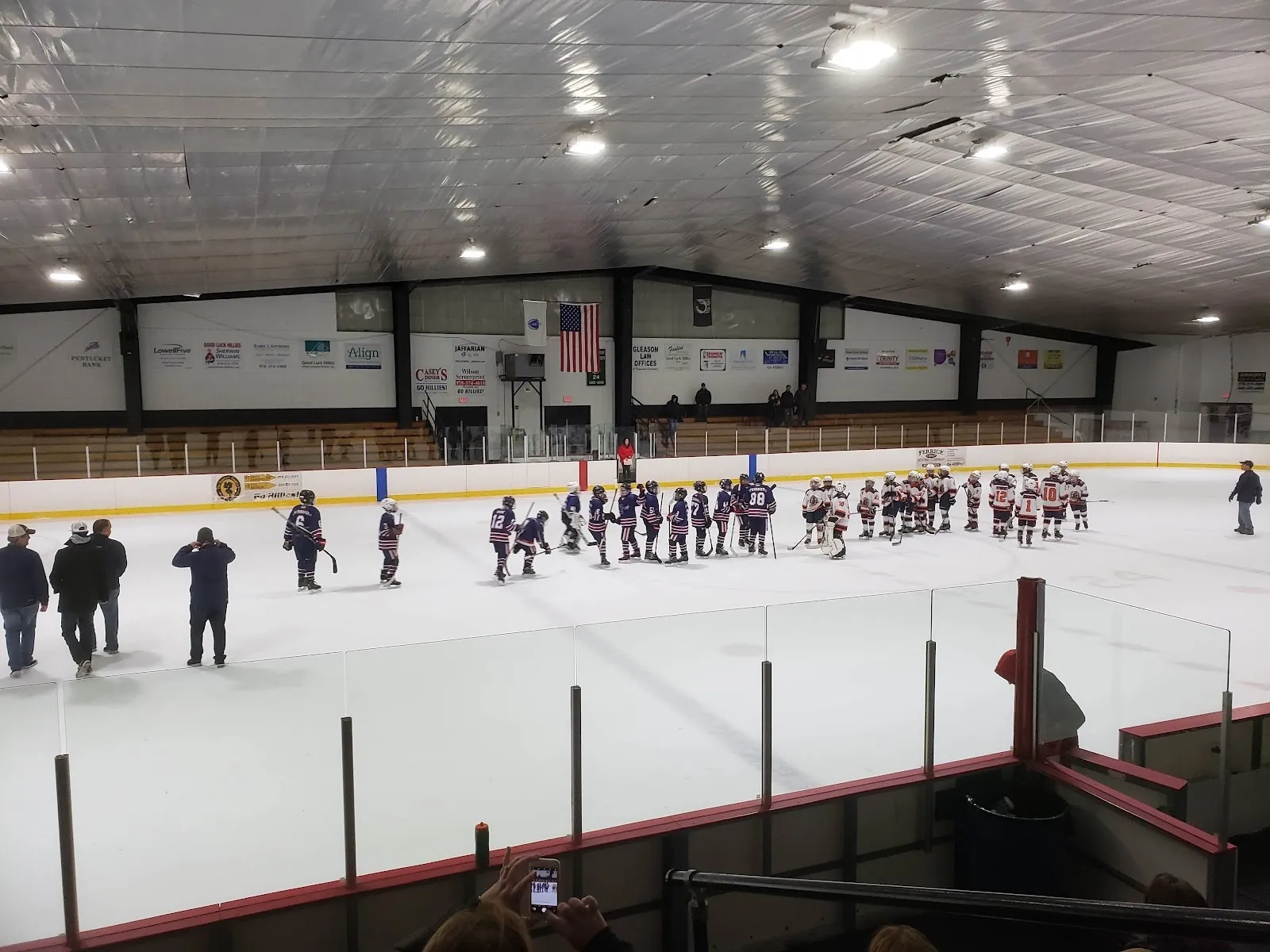 Una fila de jóvenes jugadores a lo ancho de la pista antes de un partido, vista por encima de las vallas, bajo un techo oscuro con banderines de patrocinadores en la pared del fondo. — Haverhill Veterans Memorial Rink