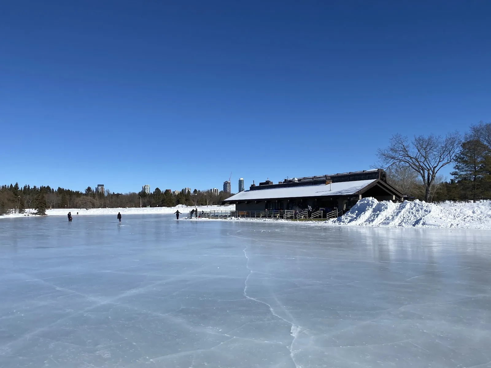 Vista amplia del hielo de la laguna congelada de Hawrelak Park, con el pabellón del parque al otro lado de la superficie y algunos patinadores a lo lejos — Hawrelak Park Outdoor Ice Rinks