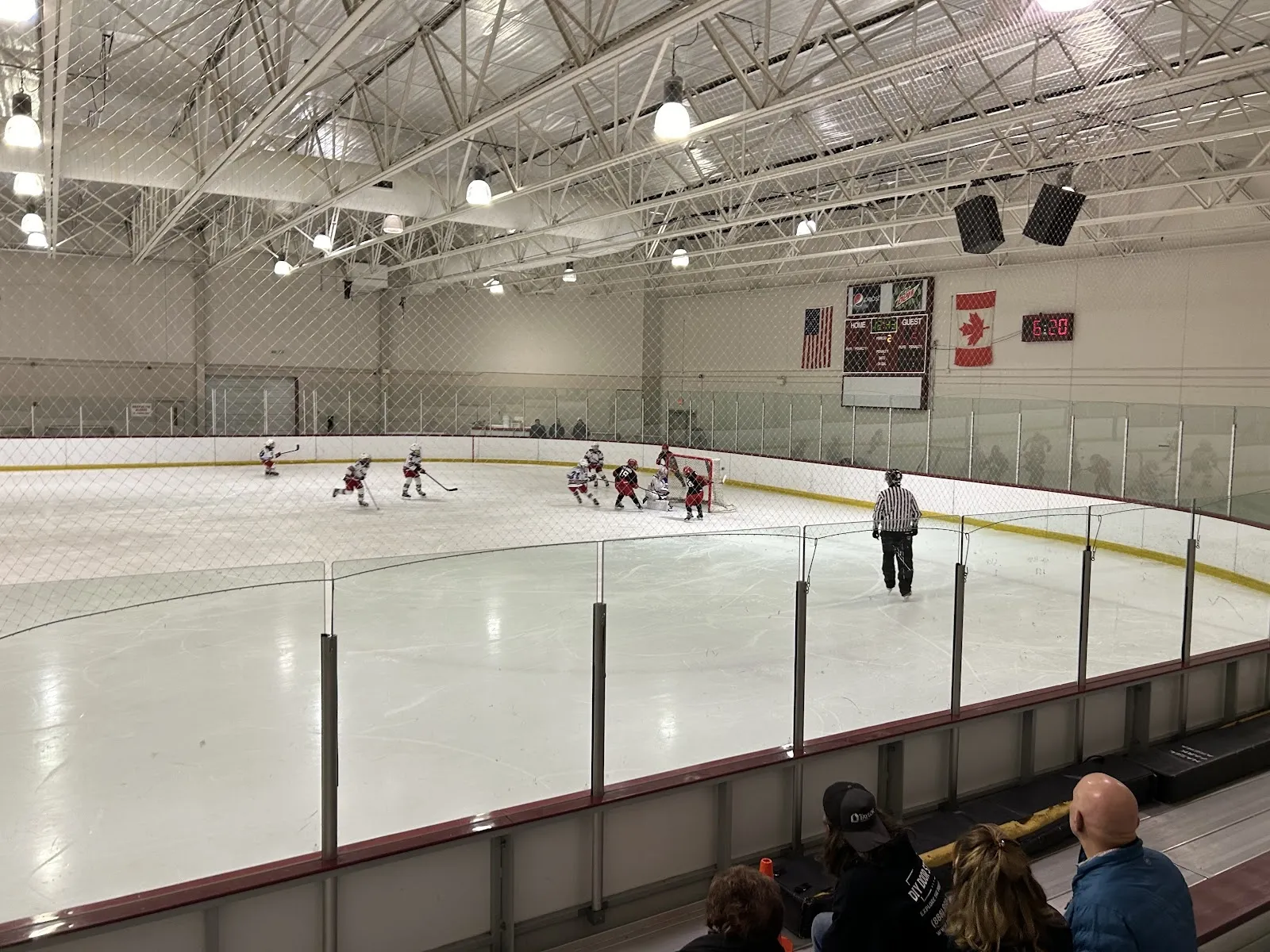 Un partido visto por encima del vidrio en las vallas, con jugadores repartidos por una pista amplia bajo un techo de acero claro, con un reloj de marcador en la pared del fondo. — Hazel Park Ice Arena