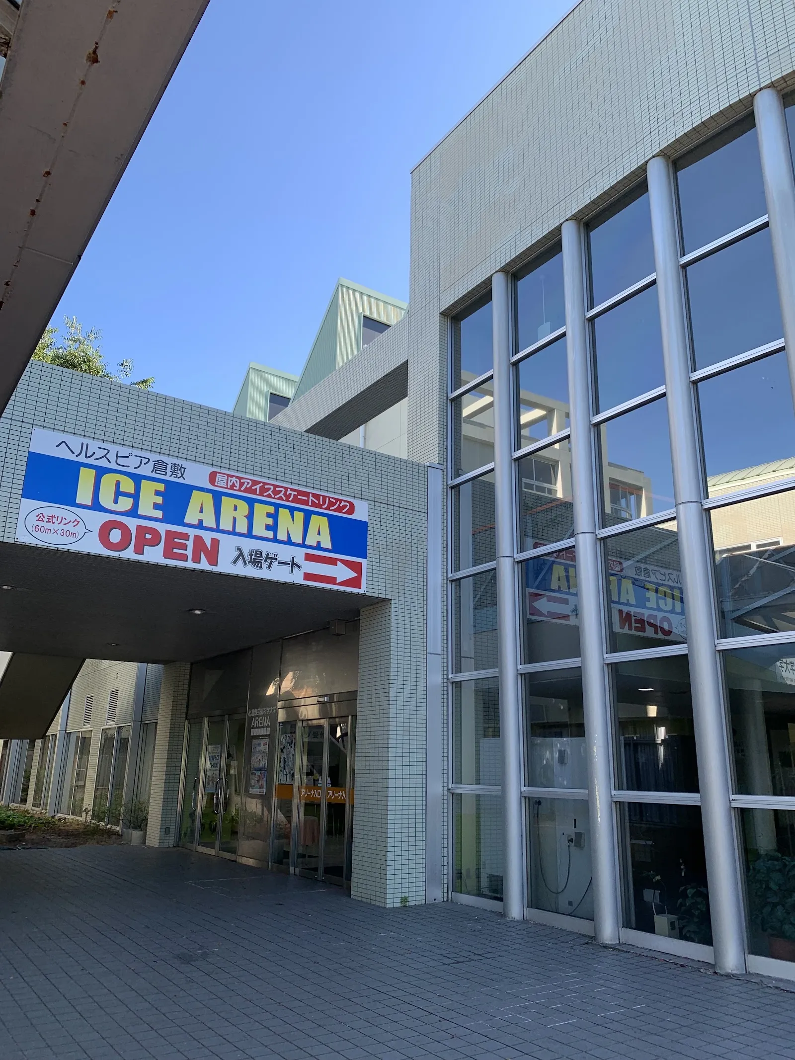 背の高いガラスのファサードのそばにある、青い「ICE ARENA OPEN」の看板の下の、ヘルスピア倉敷アイスアリーナの入口 — Healthpia Kurashiki Ice Arena
