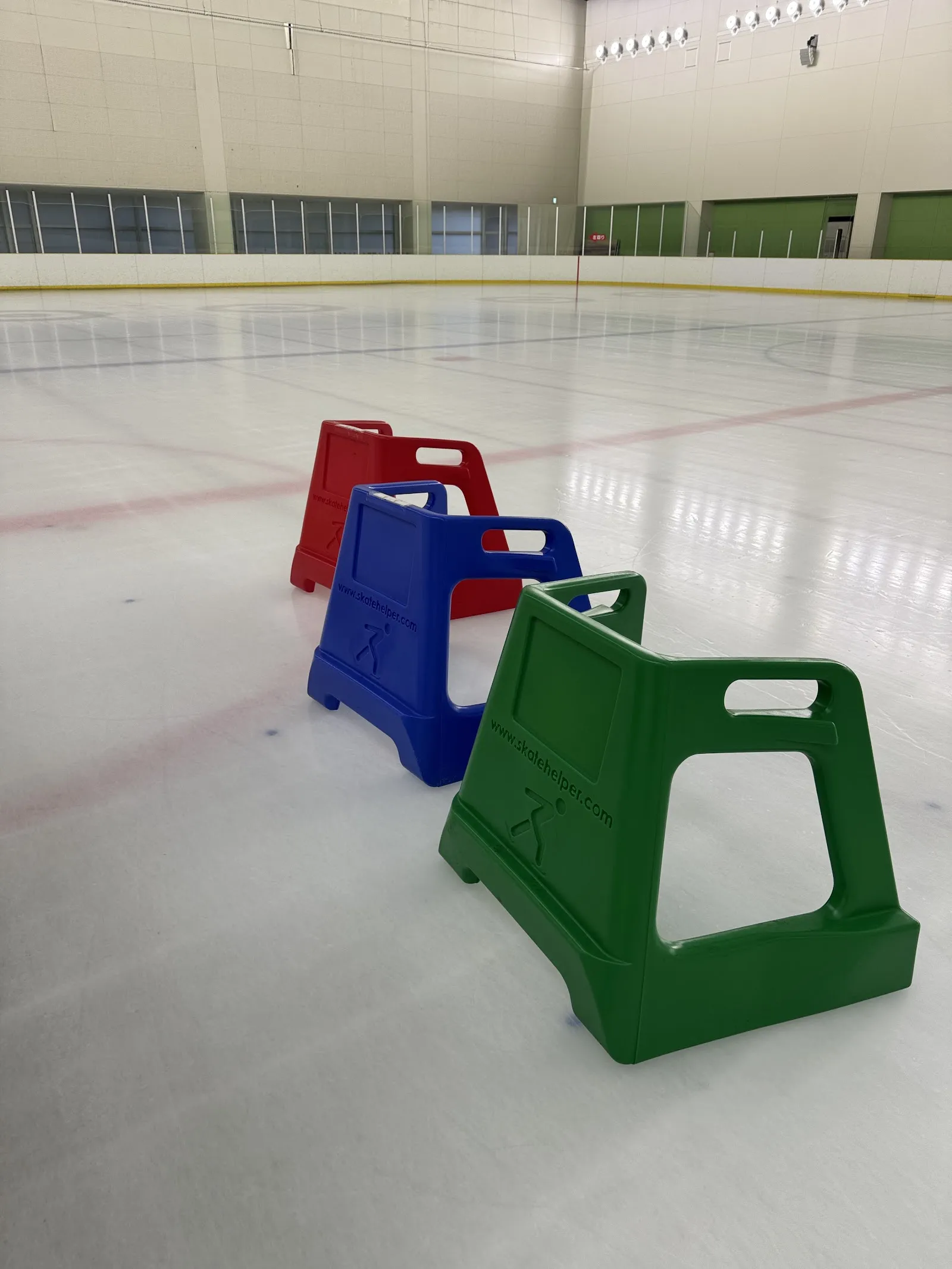ヘルスピア倉敷アイスアリーナの無人の氷面。氷の上に3色のスケートの補助具があり、後ろにホールの奥の壁が見えます — Healthpia Kurashiki Ice Arena