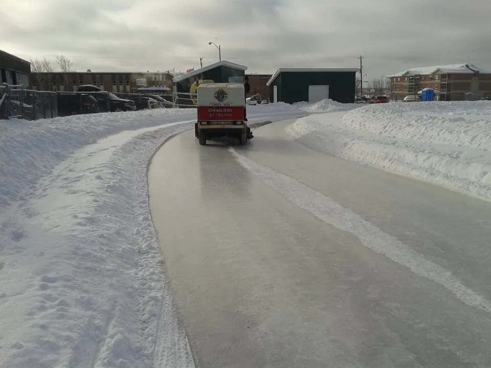 An ice resurfacing machine driving along the curved ice path of the Hearst outdoor rink between snowbanks, with town buildings behind. — Hearst Outdoor Ice Rink