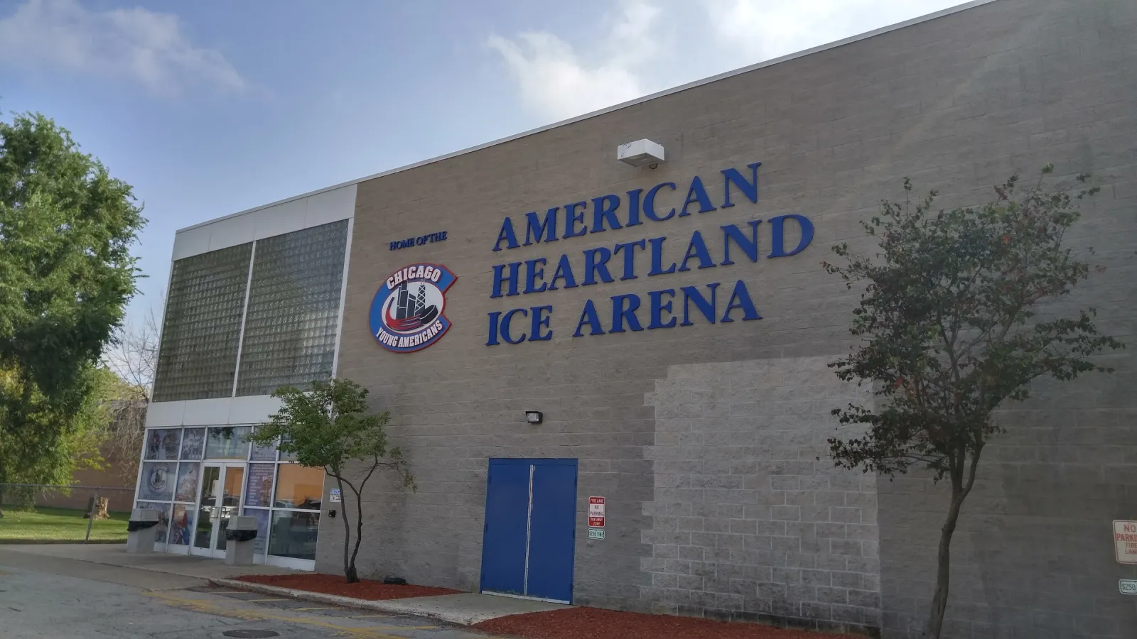 The grey block building with "American Heartland Ice Arena" in large blue letters, a logo, a blue side door and the glass entrance at left. — American Heartland Ice Arena