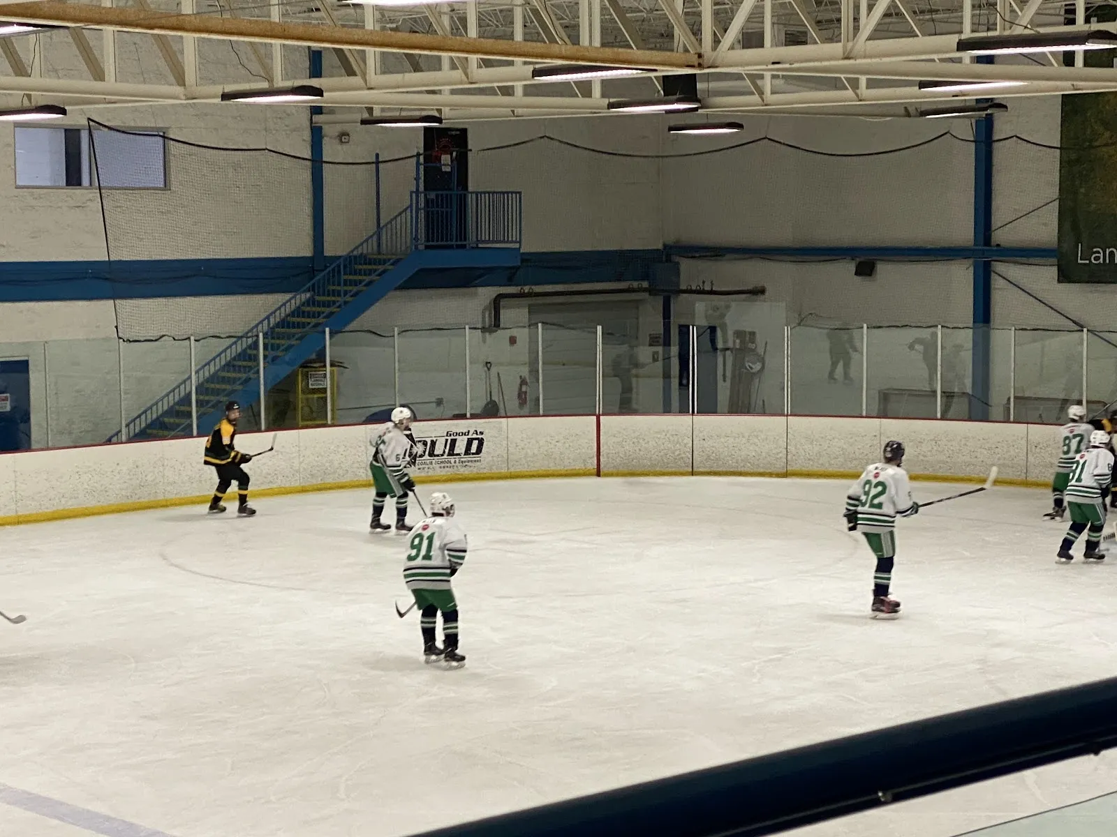 A game at one end of the sheet seen past a goal, players spread across the ice with a blue-railed stair behind them under a steel roof. — American Heartland Ice Arena