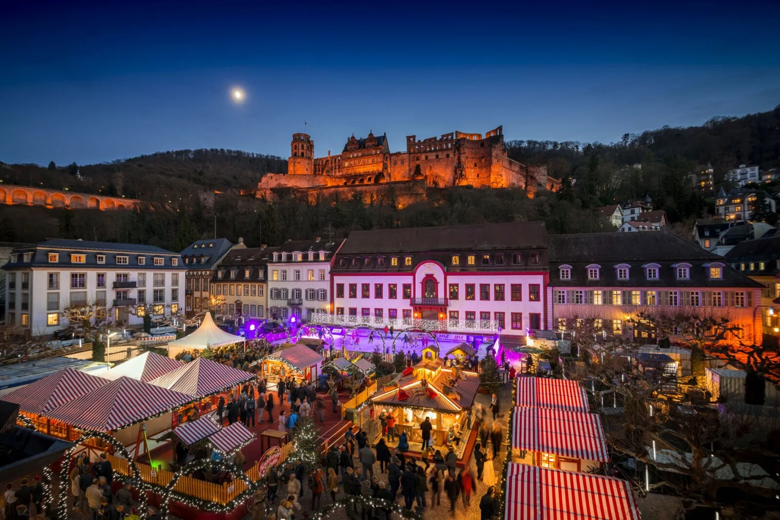 The Christmas market on the Karlsplatz at dusk with the lit rink among the stalls and the floodlit Heidelberg castle on the hill above the old town. — Heidelberger Eisbahn