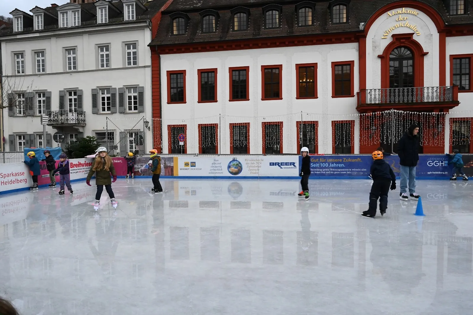 The open-air rink on the Karlsplatz with its sponsor boards, a few skaters on the pale ice and the white and red façade of the Akademie der Wissenschaften behind. — Heidelberger Eisbahn