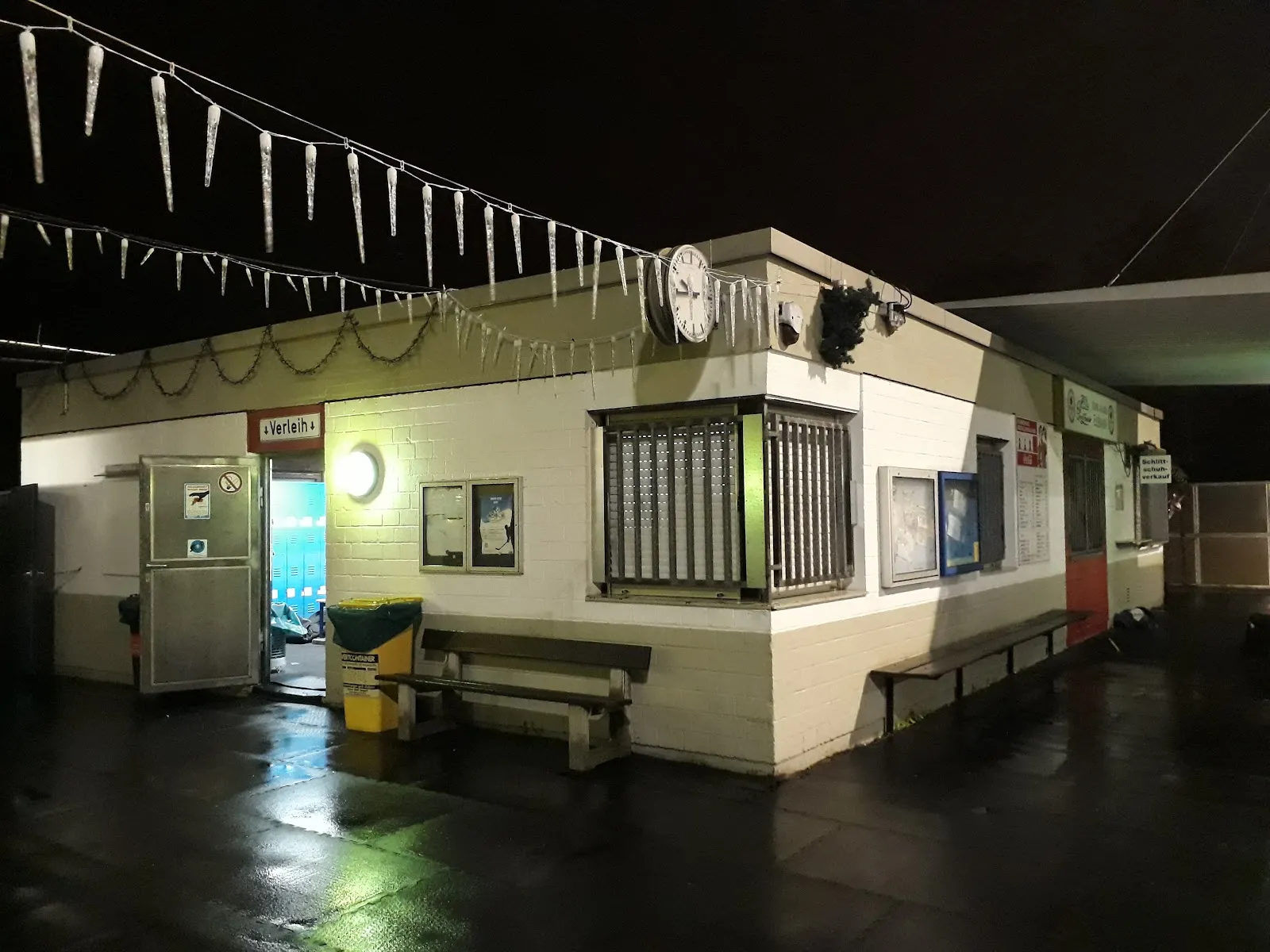 Das kleine Eingangsgebäude der Freiluft-Eisbahn bei Nacht, mit einer Uhr an der Wand, Eiszapfen-Lichterketten entlang der Traufe und offener Eingangstür. – Henkell-Kunsteisbahn