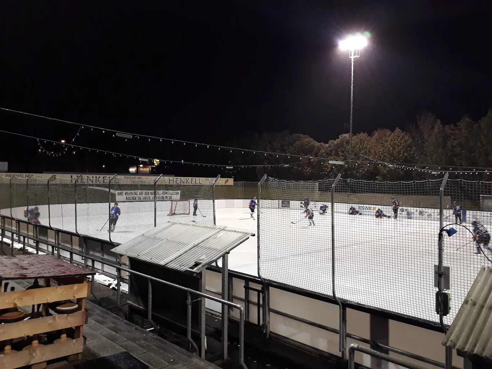Die Freiluft-Eisbahn in der Dämmerung, ihre Fläche von Flutlicht beleuchtet, mit kleinen Eisläufern in der Ferne sowie Banden und Zaun ringsum. – Henkell-Kunsteisbahn