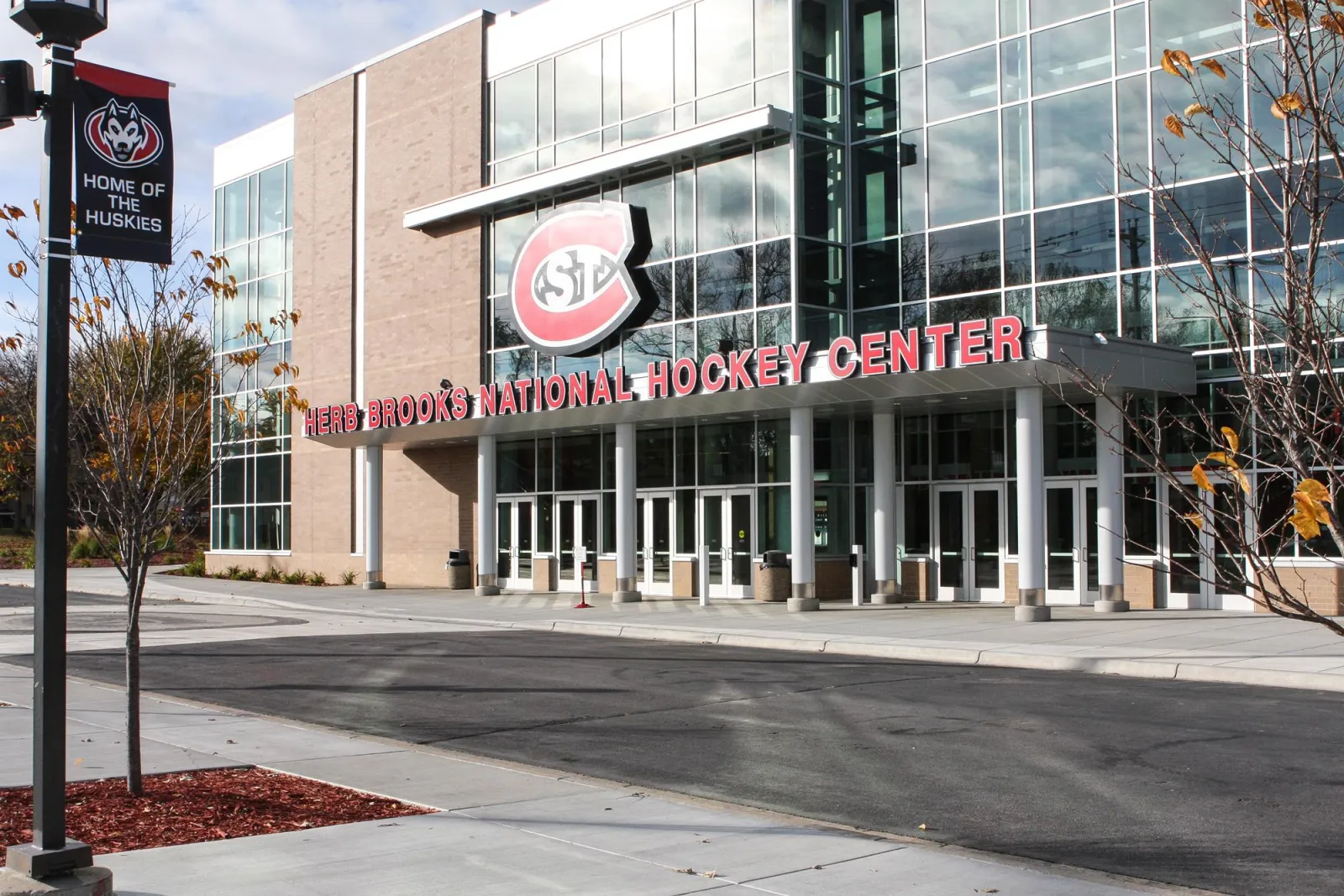 The glass entrance atrium with the St. Cloud State logo and HERB BROOKS NATIONAL HOCKEY CENTER lettering, a Huskies banner post on the left. — Herb Brooks National Hockey Center