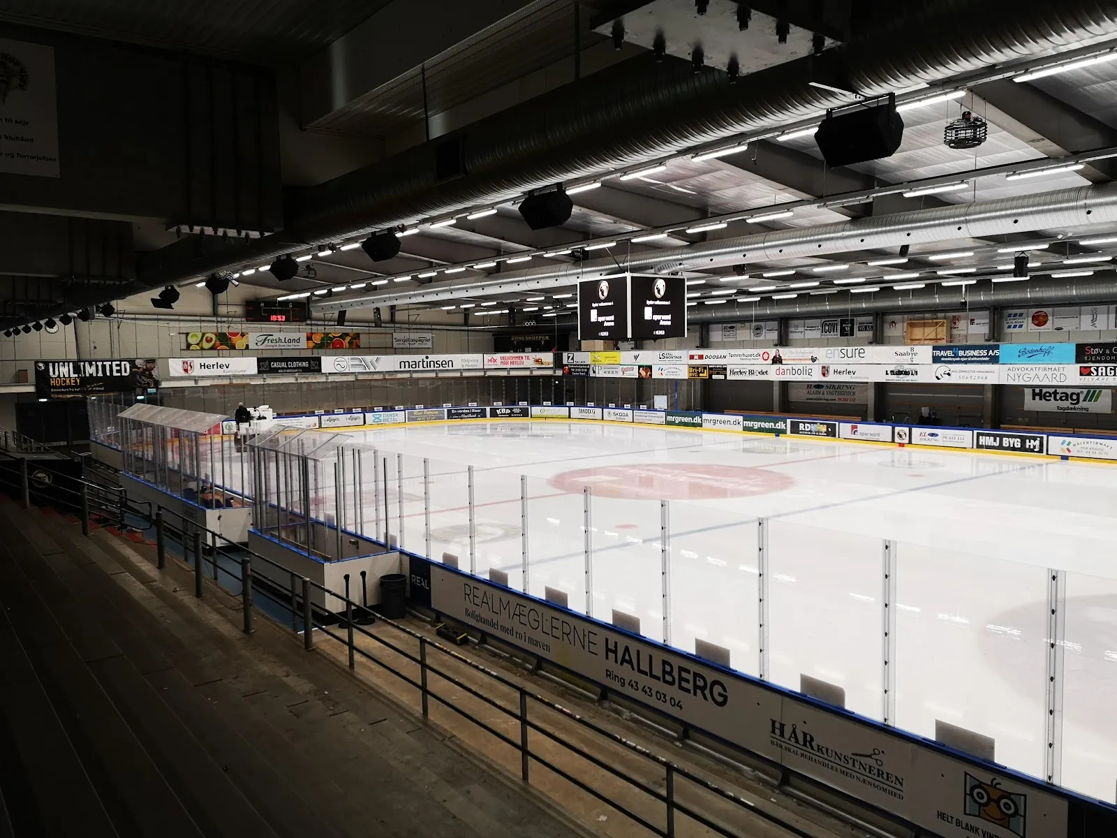 The empty ice sheet of Herlev Skøjtehal seen from the upper stands, with the rink boards, scoreboards and a long row of sponsor panels along the far side. — Herlev Skøjtehal