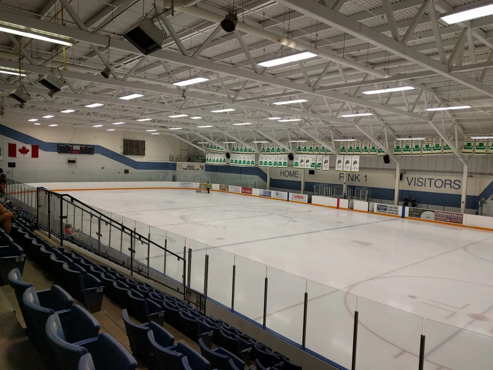 The empty ice at Hespeler Memorial Arena from the stands, with blue seating, the Rink 1 and Home and Visitors signs and banners along the far wall — Hespeler Memorial Arena