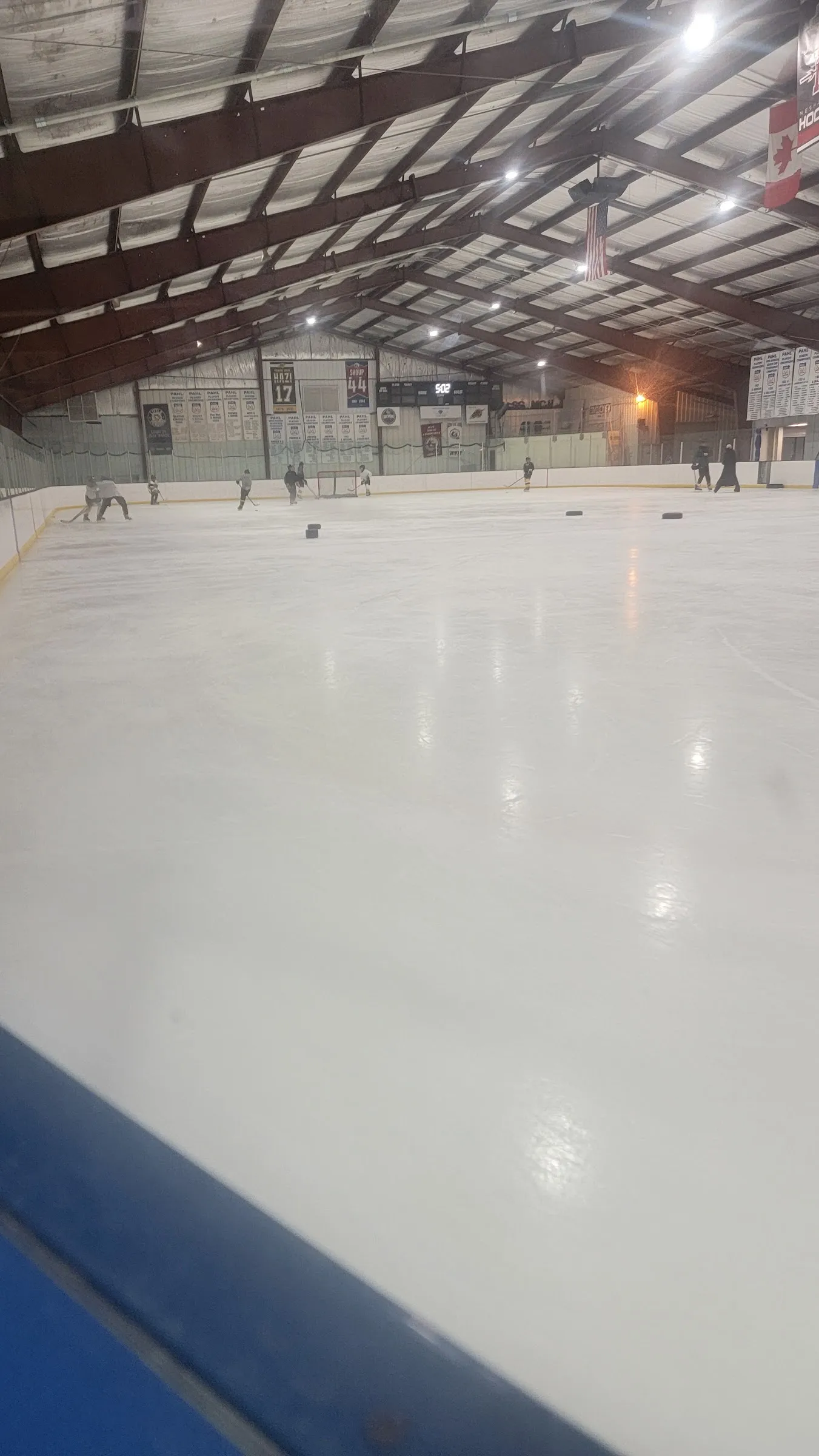 El hielo visto desde un extremo, con algunos patinadores cerca de las vallas del fondo, una bandera de Canadá y mantas bajo vigas rojo oscuro en el techo. — Hess Ice Rink