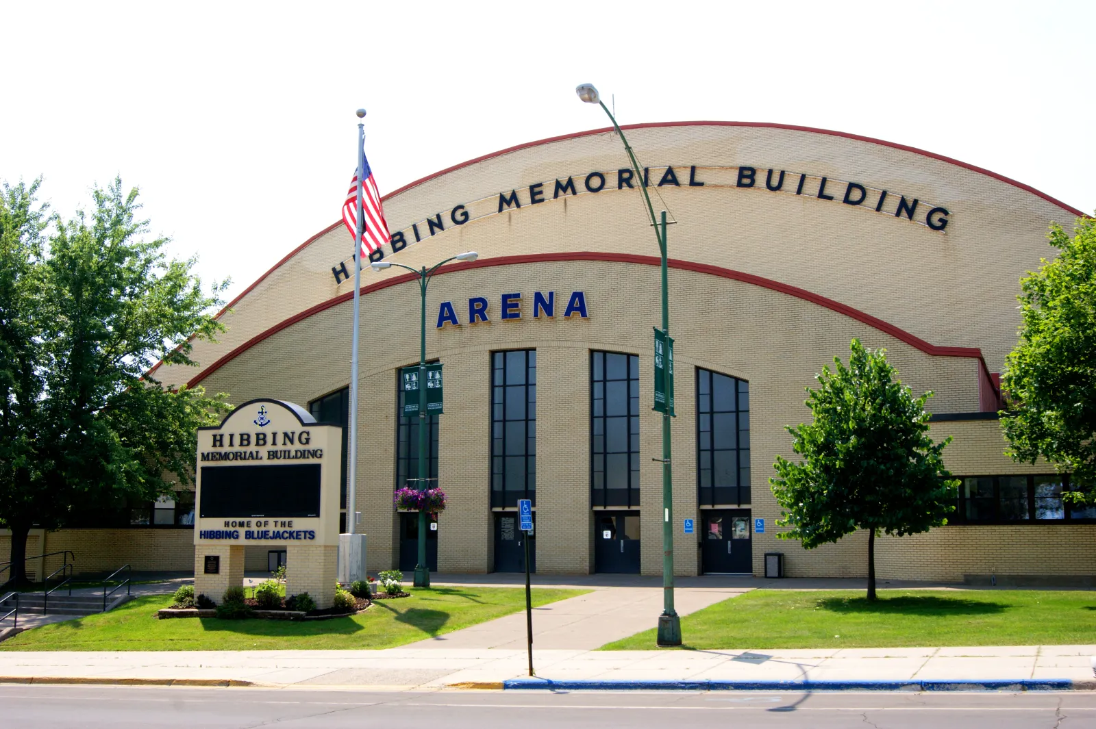 La fachada arqueada del Hibbing Memorial Building, con las letras HIBBING MEMORIAL BUILDING y ARENA sobre altas ventanas, el letrero 'Home of the Hibbing Bluejackets' y un asta de bandera en el césped. — Hibbing Memorial Building Arena