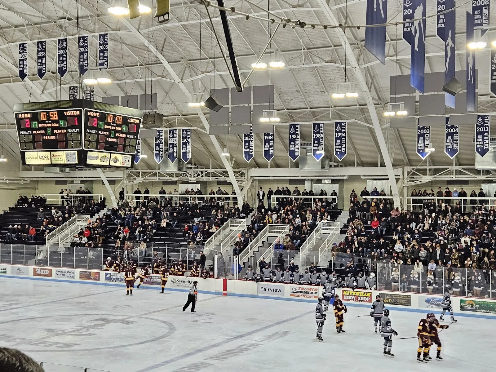 Un partido visto desde las butacas bajas, con jugadores repartidos por el hielo debajo de un techo abovedado de la arena con banderines azul marino y un marcador sobre las gradas. — Hibbing Memorial Building Arena