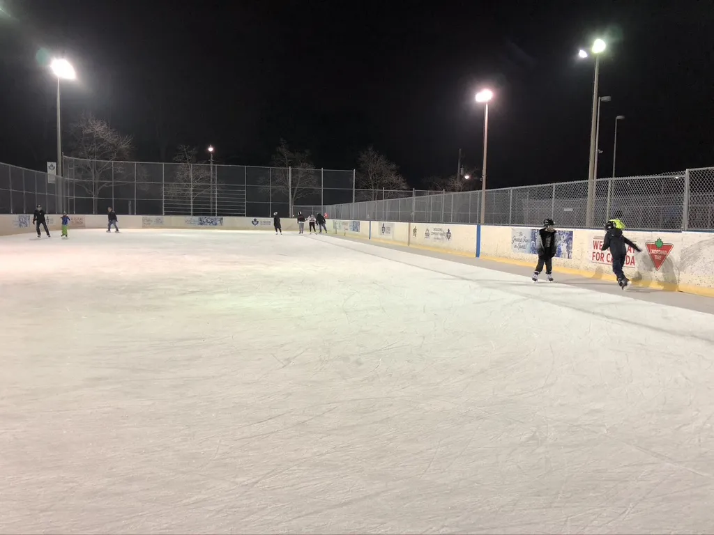 La superficie de hielo de la pista al aire libre de High Park de noche en invierno, con bardas, cercas y reflectores, y algunos patinadores repartidos por el hielo — High Park Outdoor Ice Rinks