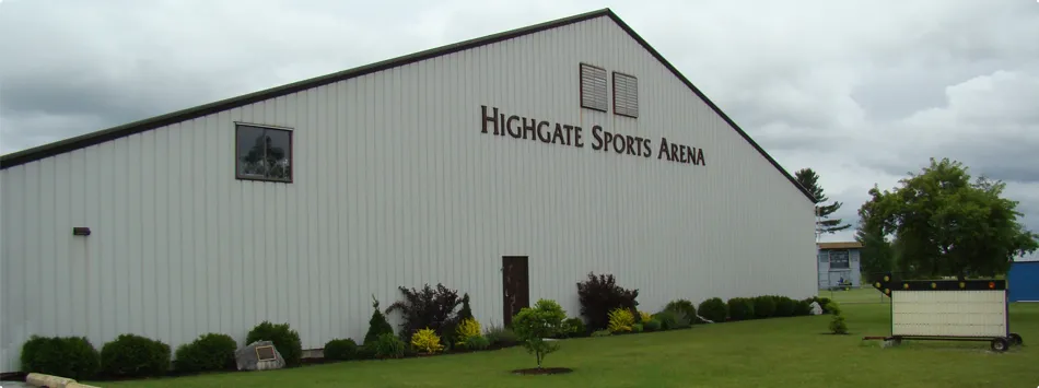 Side and gable end of the metal-sided Highgate Sports Arena building, with a 'HIGHGATE SPORTS ARENA' sign on the gable and landscaped shrubs along the entrance. — Highgate Sports Arena