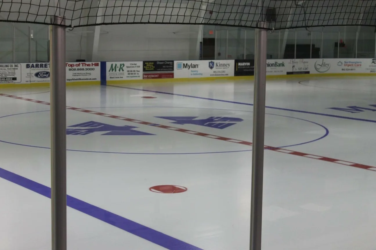 Wide view of the empty ice sheet through a goal net, with local-sponsor dasherboard ads (including a Highgate, VT business) lining the boards. — Highgate Sports Arena