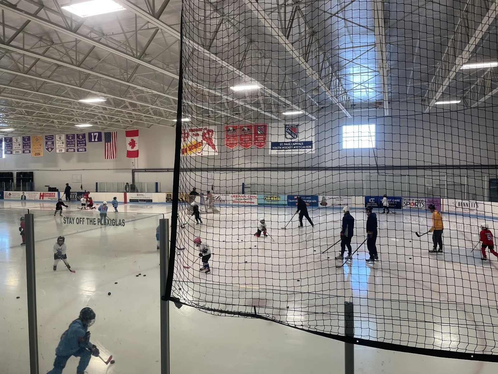 Children skating and a few adults spread across the sheet seen through a black net, flags and banners hung along the far wall under steel trusses. — Charles M. Schulz–Highland Arena