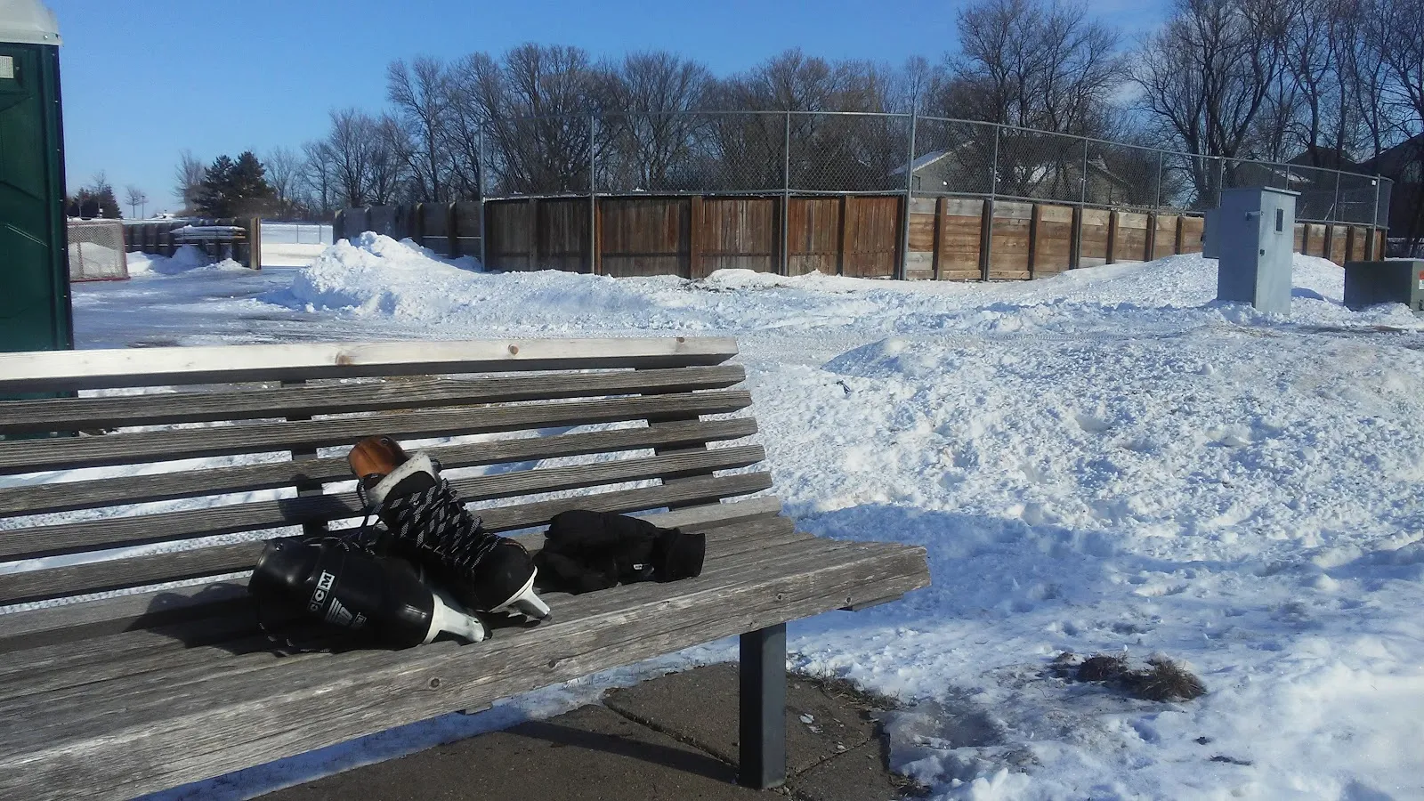 A bench with a pair of skates and a bag beside the rink's wooden boards at Highview Heights Park, with snow banks, a utility box and bare trees behind in winter sun. — Highview Heights Park Ice Rinks