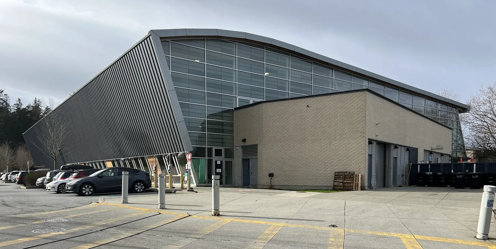 The Hillcrest Rink building in Vancouver, with its slanted metal wall, glass curtain wall and brick service block, seen from the parking lot — Hillcrest Rink