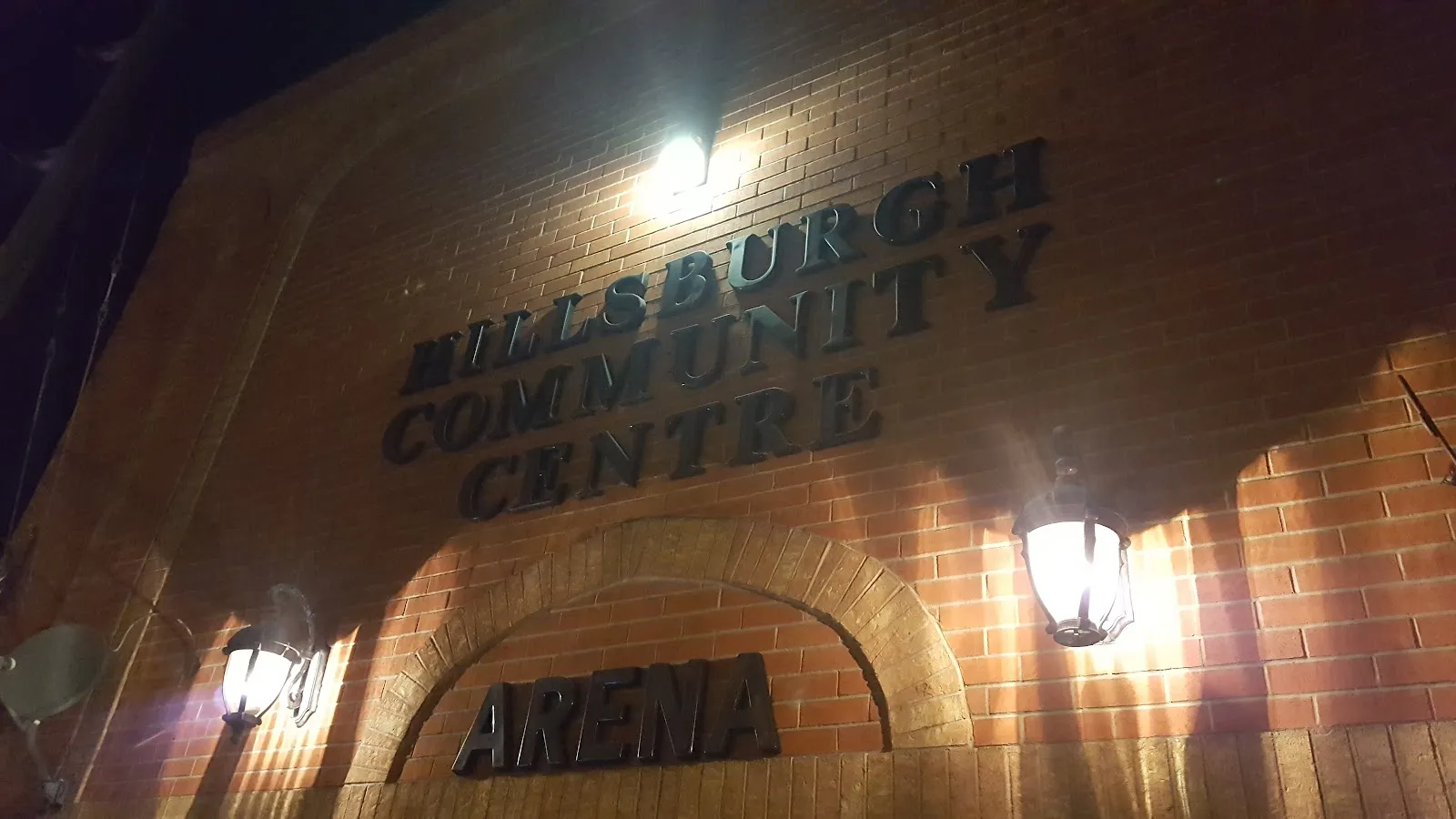The brick front of the Hillsburgh Community Centre at night, with its name carved above an arch marked Arena and two wall lamps lighting the entrance. — Hillsburgh Community Centre