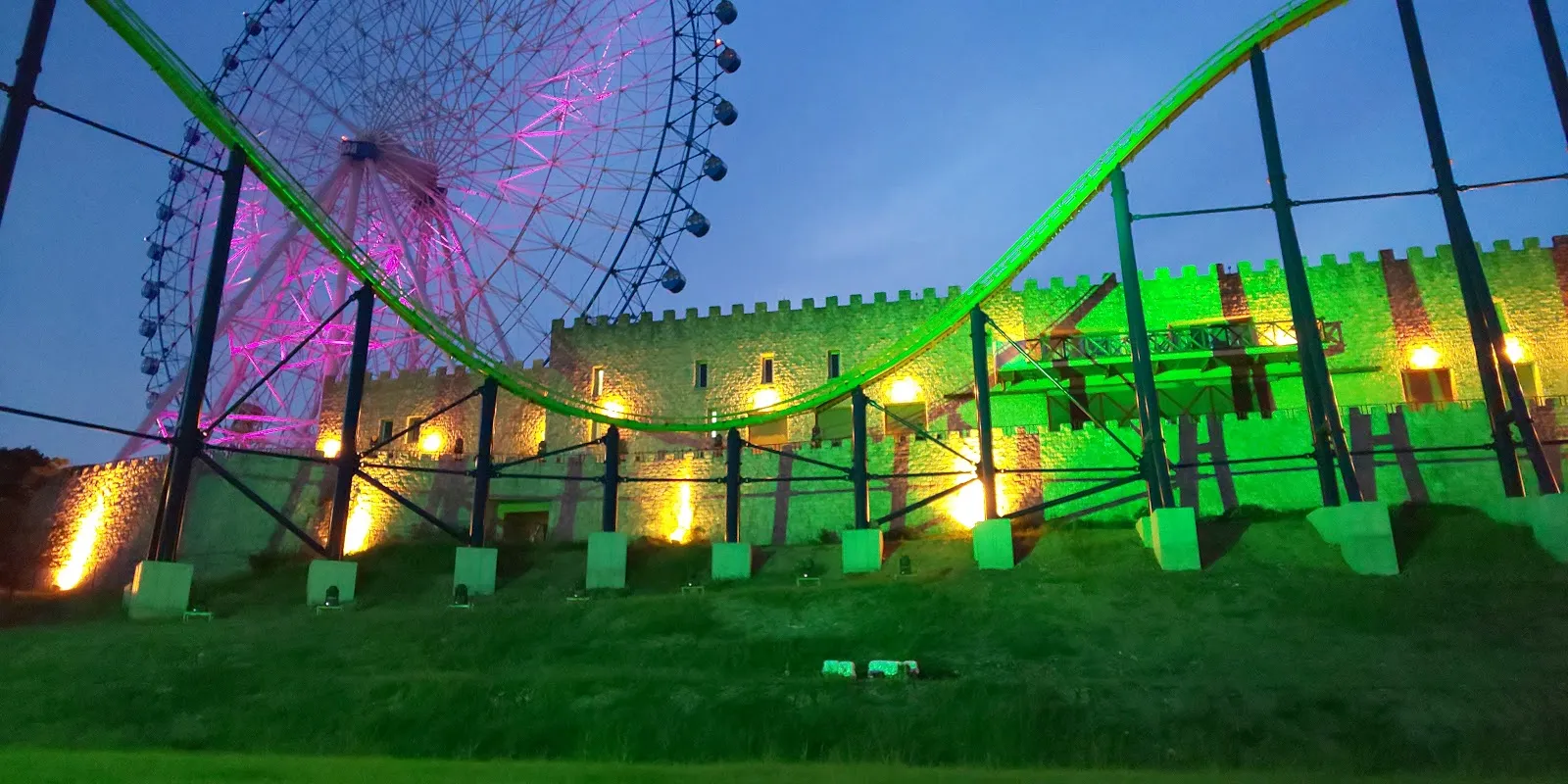 The castle-like hall of Himeji Central Park at dusk, with a Ferris wheel lit pink behind it and a roller-coaster track lit green in front. — Himeji Central Park Ice Park