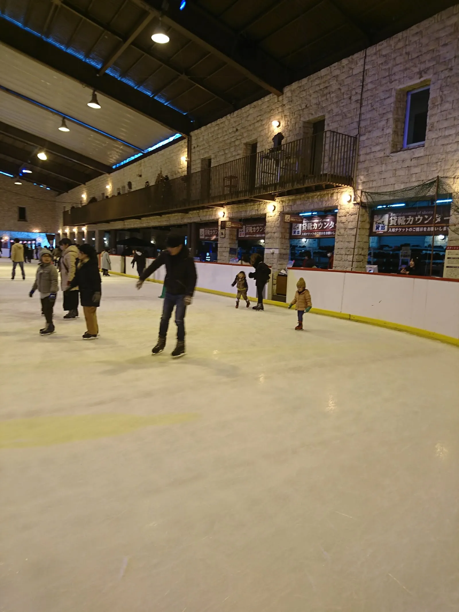 Skaters on the indoor ice at Himeji Central Park, beside the rental counters set into a stone-faced wall under a wooden roof. — Himeji Central Park Ice Park