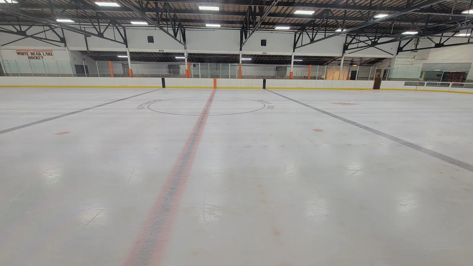 The empty sheet from rinkside, with the red centre line running to the far boards, the White Bear Lake Hockey banner and the steel roof trusses above. — Hippodrome Ice Arena