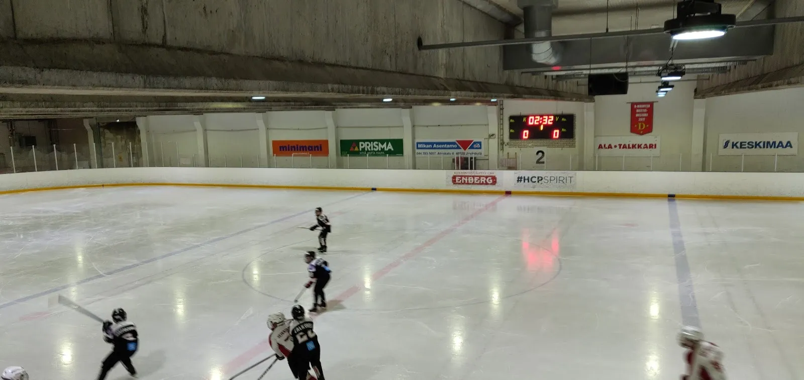 A skater on the ice of the Hippos practice rink in Jyväskylä, seen across the sheet with the boards and the hall roof beyond. — Hippoksen harjoitusjäähalli