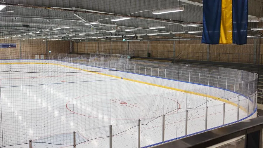 Den tomma isen i HLL Arena i Nynäshamn sedd från läktaren genom skyddsnätet, med sargen och en träklädd vägg bakom – HLL Arena (Nynäshamn)