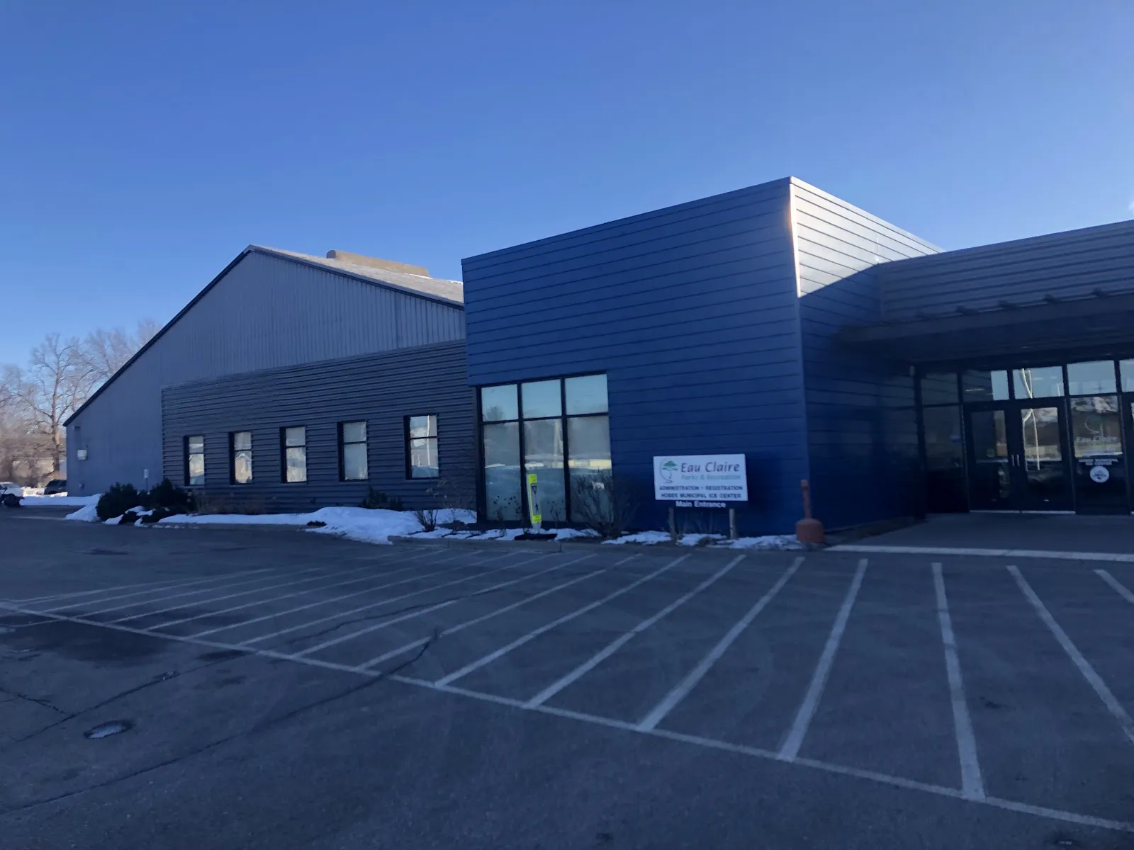 The blue-clad entrance block and arena hall seen from the parking lot, with the City of Eau Claire sign by the doors. — Hobbs Ice Arena