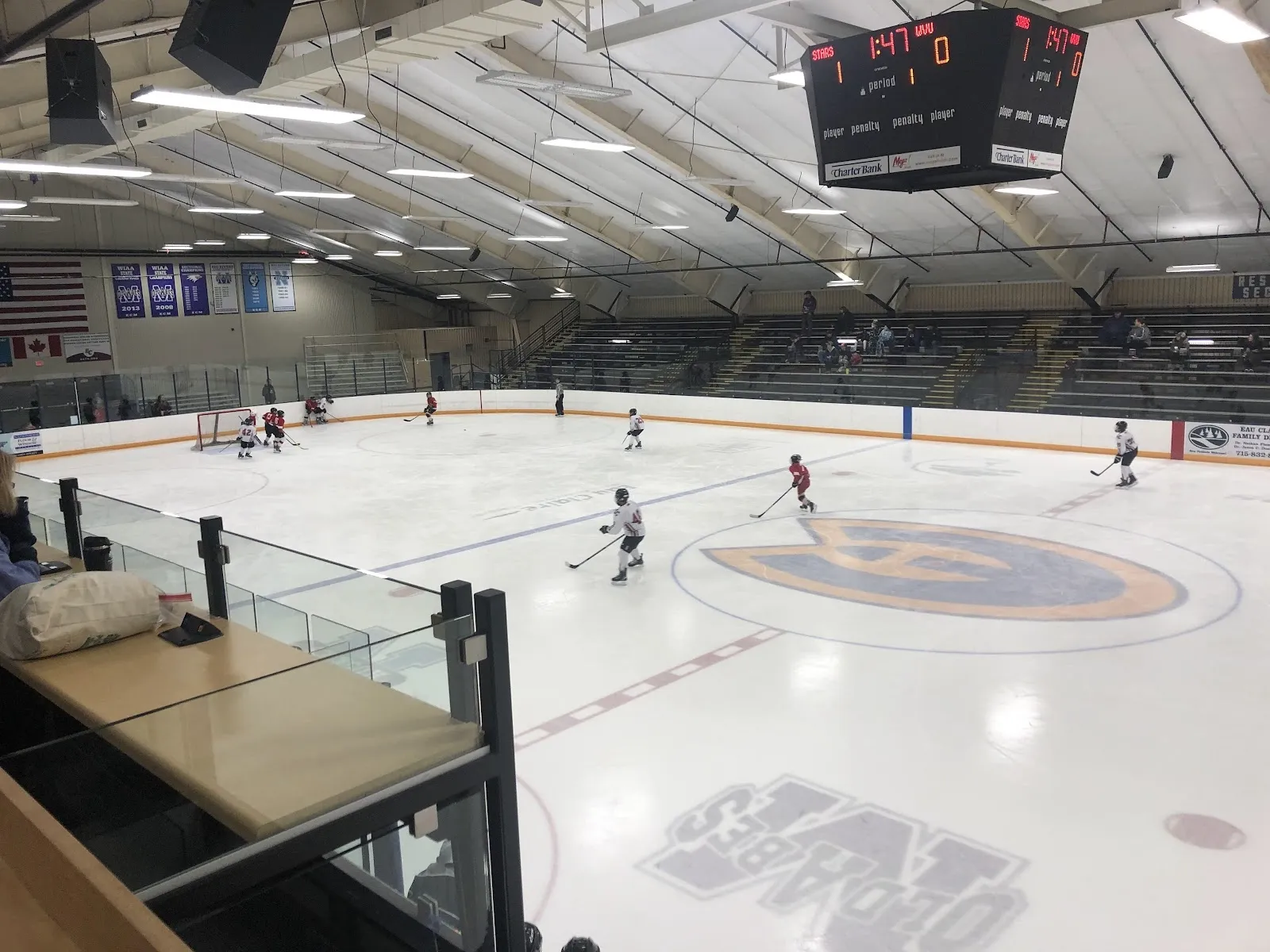 A wide view from beside the benches, the sheet with a university logo at centre circle under a scoreboard and a steel-trussed ceiling. — Hobbs Ice Arena
