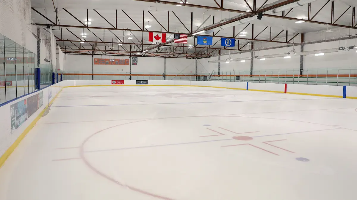 La pista vacía de Hockey Hounds desde el centro del hielo, con banderas de Canadá, Estados Unidos y Alberta bajo armaduras rojas del techo. — Hockey Hounds Recreation Centre