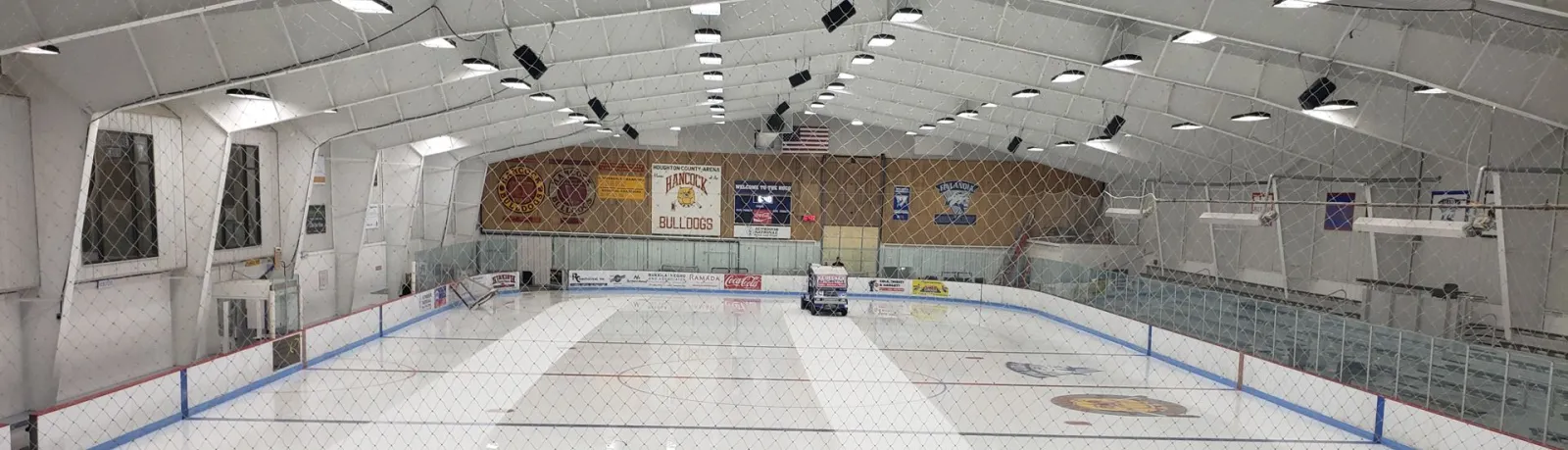 Wide view of the empty HoCo Arena sheet under a white arched roof, with blue-trimmed boards and glass, team banners on the far wall and bleachers along the side. — HoCo Arena