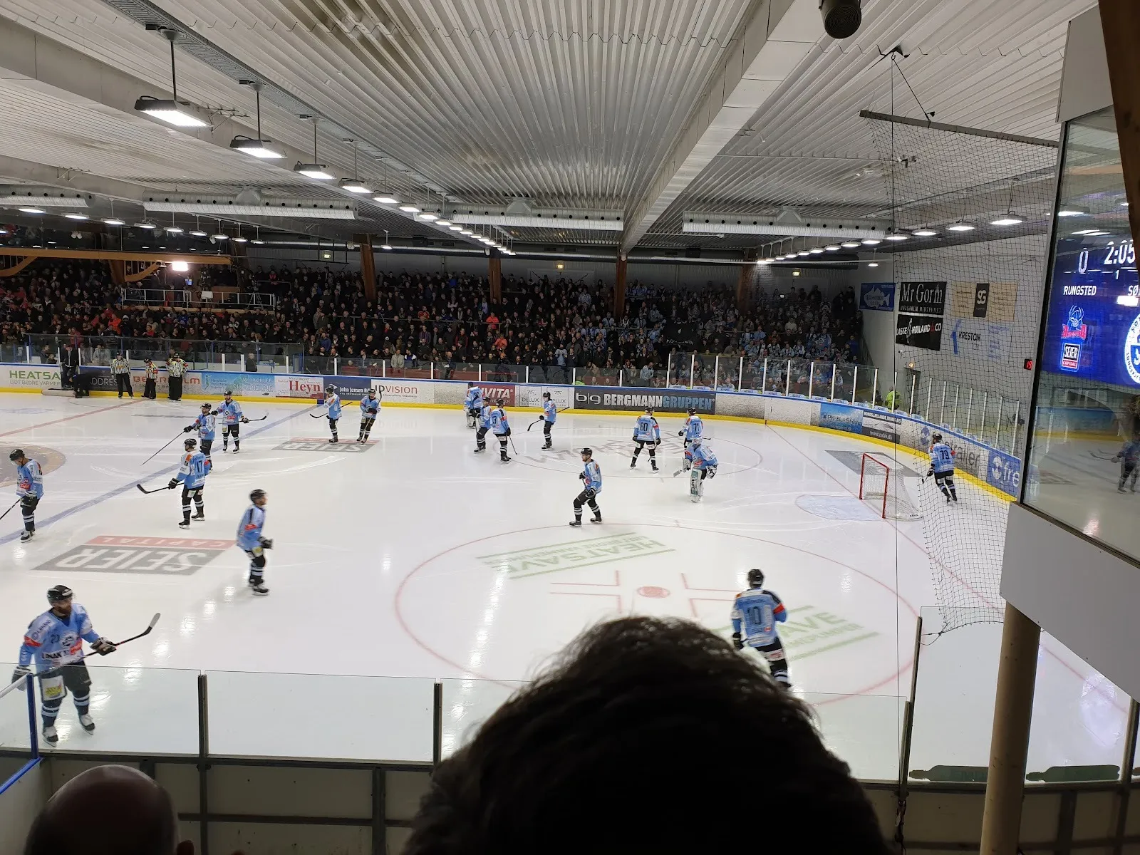 A game on the ice at the Hørsholm rink seen from behind the glass, players across the rink with spectators and sponsor boards around it. — Hørsholm Skøjtehal