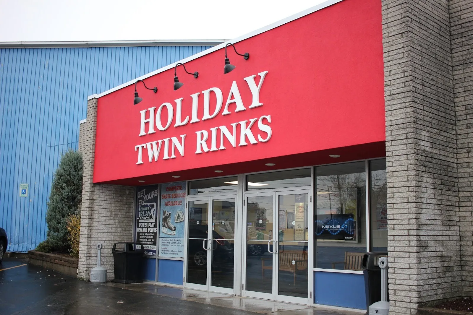 La entrada de Holiday Twin Rinks, con su nombre en letras blancas sobre una franja roja encima de puertas de vidrio, junto a la nave azul de la pista — Holiday Twin Rinks