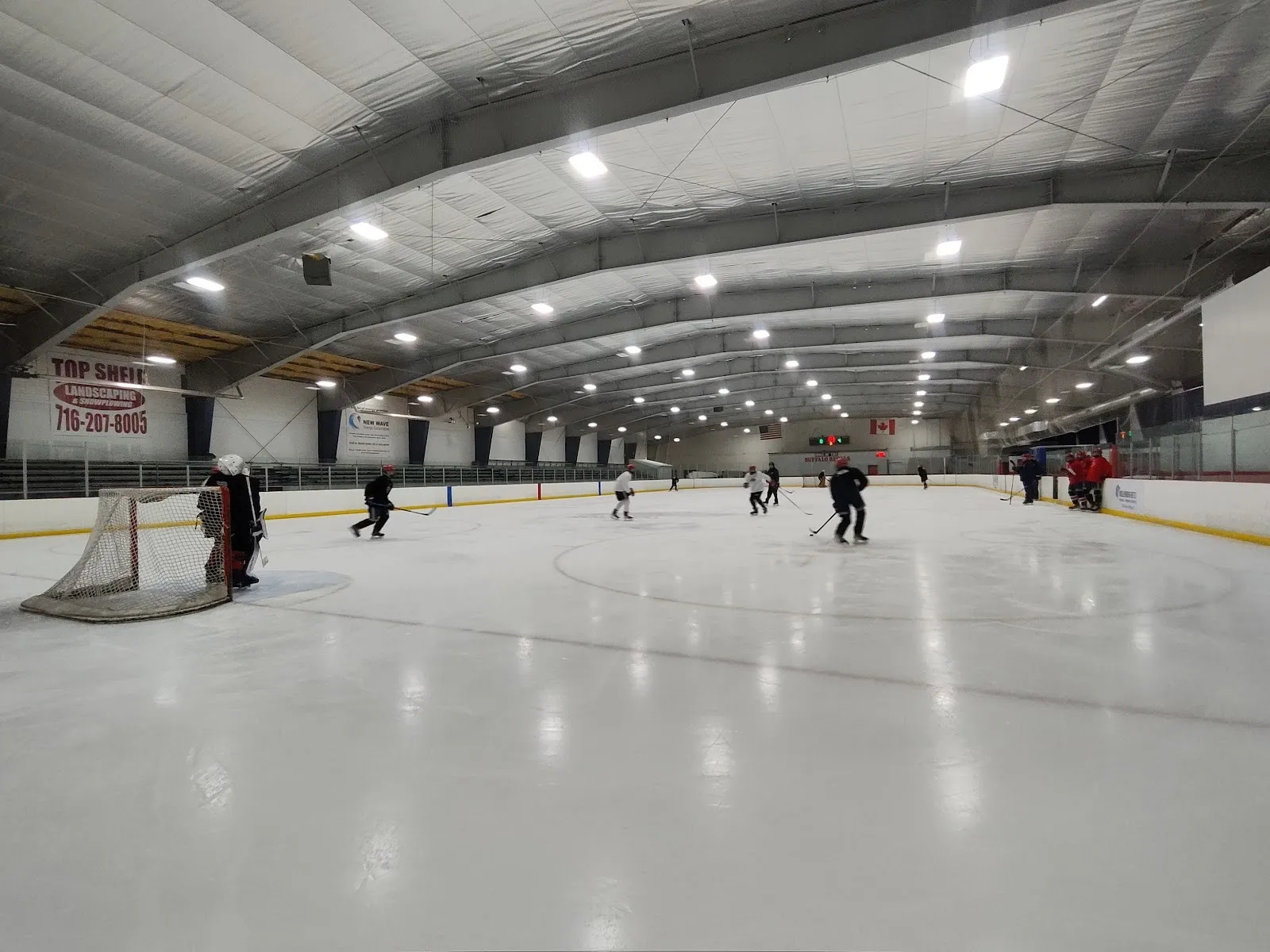 Una vista larga a lo largo de la pista desde las vallas cercanas, con unos cuantos jugadores repartidos a lo lejos por el hielo bajo un techo curvo de acero iluminado con hileras de luces. — Holiday Twin Rinks