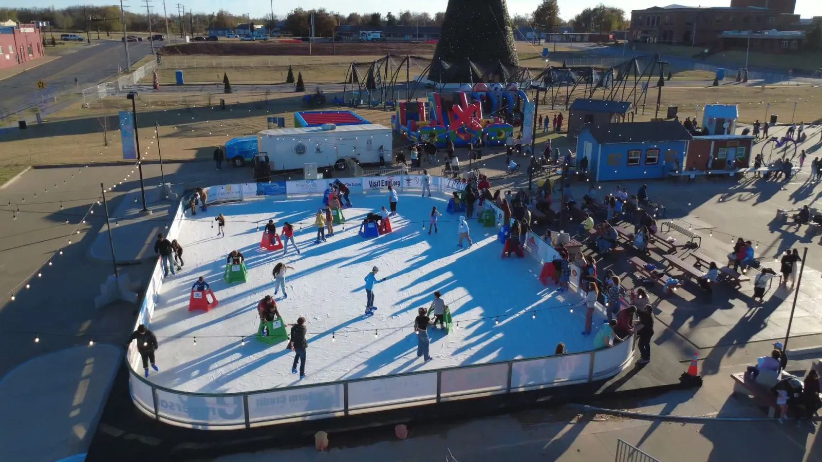 Aerial view of the Downtown Enid ice rink set up in a parking lot, with skaters on the ice, the rink's coloured huts and a play area beside it. — Holidays on Ice Downtown Enid Ice Rink