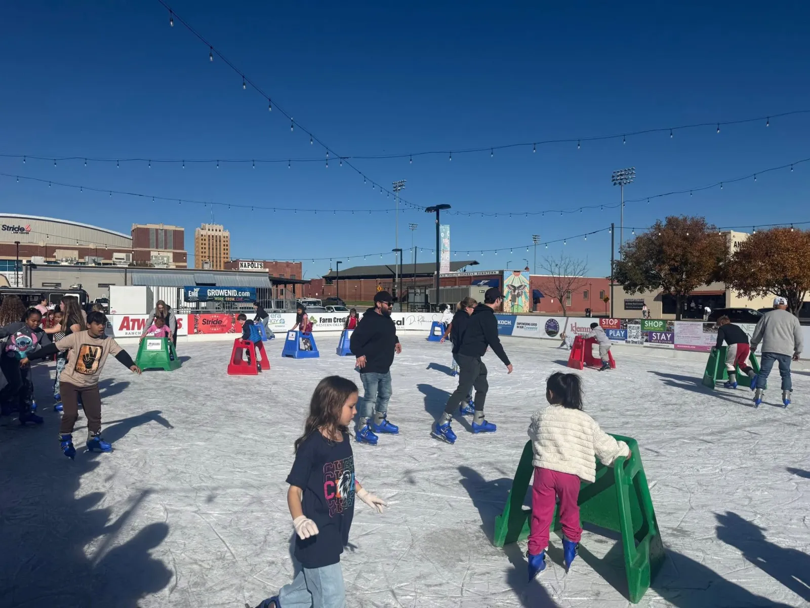 Children and adults skating on the open-air rink on a sunny day, with skate aids on the ice, sponsor boards and the downtown skyline behind. — Holidays on Ice Downtown Enid Ice Rink