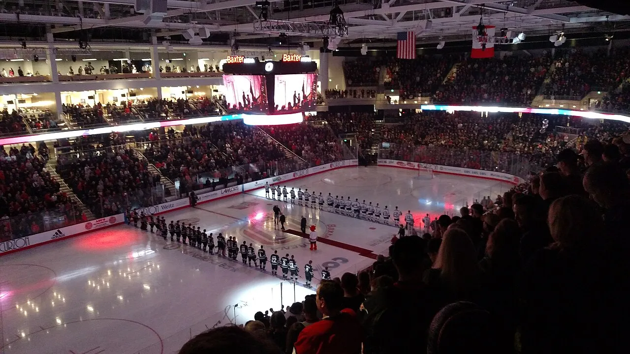 The main ice at Baxter Arena on opening night, with both teams lined up on the blue lines under the video board and a full crowd around the boards. — Holland Ice at Baxter Arena
