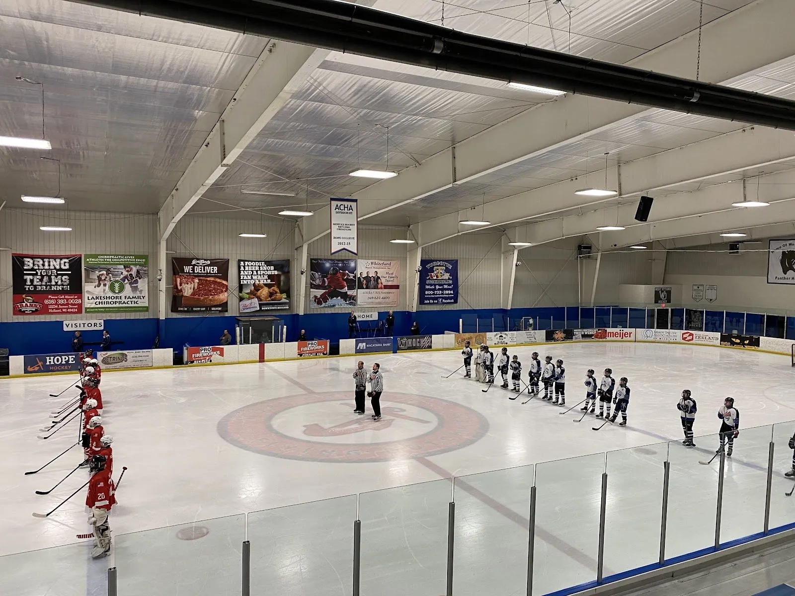 Una pista amplia vista desde las gradas, con jugadores repartidos a lo largo de las vallas del fondo bajo un techo de acero claro con franjas de banderines rojos a lo largo de la pared. — Holland Ice Arena