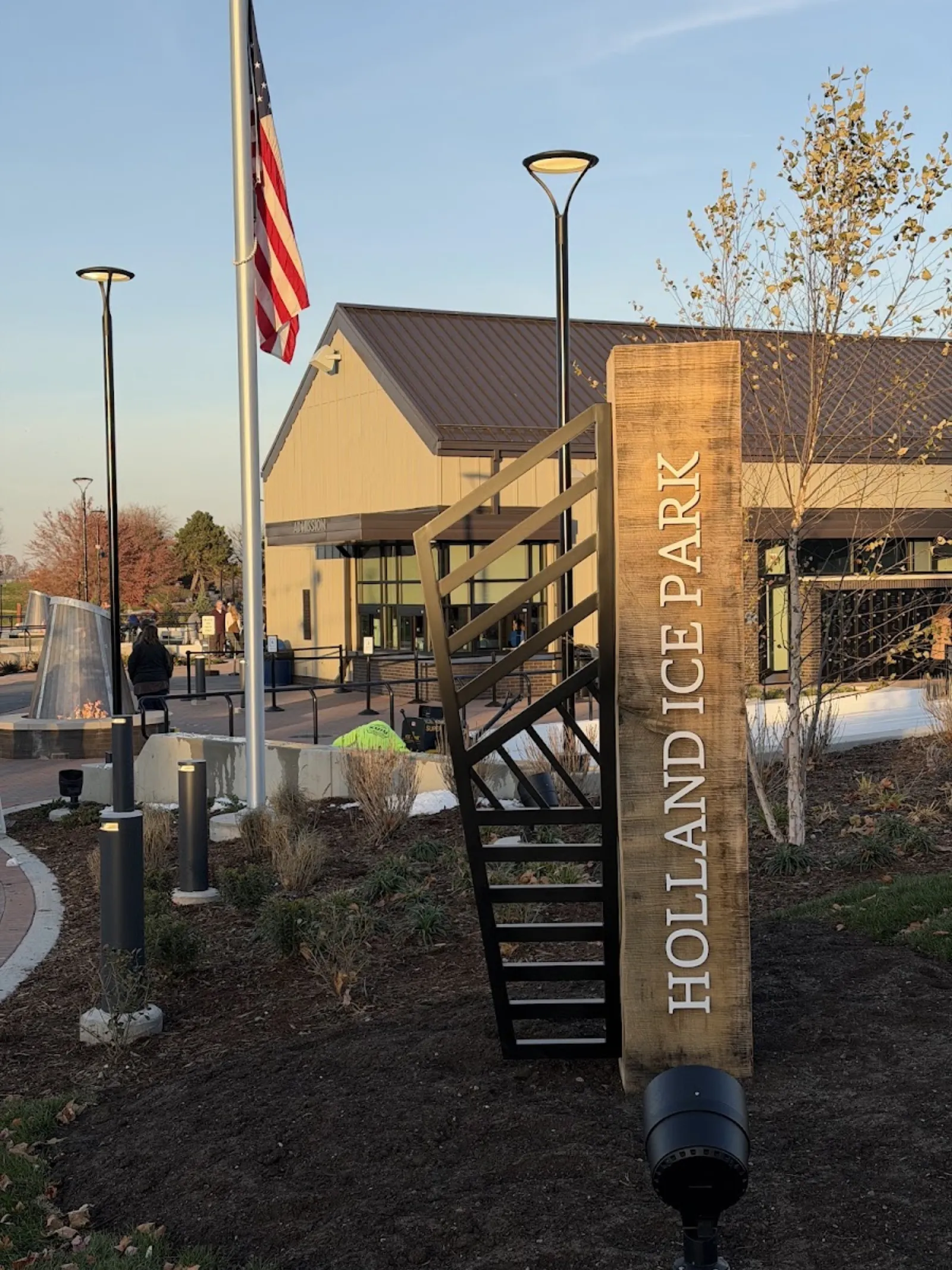 The Holland Ice Park sign and flag beside the timber admission building, with lamp posts and fire pit. — Holland Ice Park